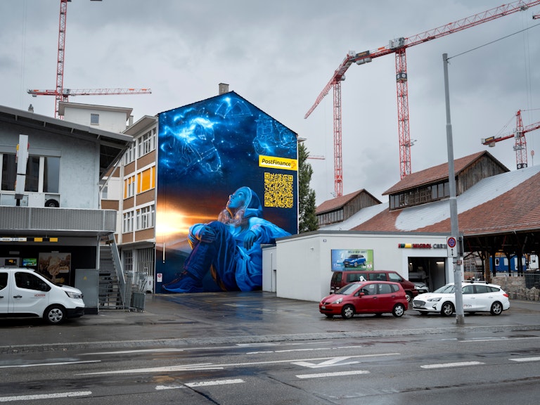 Ein Wandgemälde zeigt eine Person in Blau und kosmischer Kunst auf einem städtischen Gebäude mit Kränen, geparkten Autos und einem gelben QR-Code an der Wand.