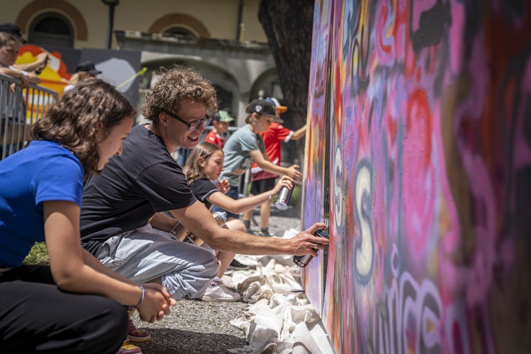 Menschen knien nieder und sprühen draußen ein lebendiges Graffiti-Wandbild, während andere in der Nähe zuschauen oder sich an der kreativen Tätigkeit beteiligen.