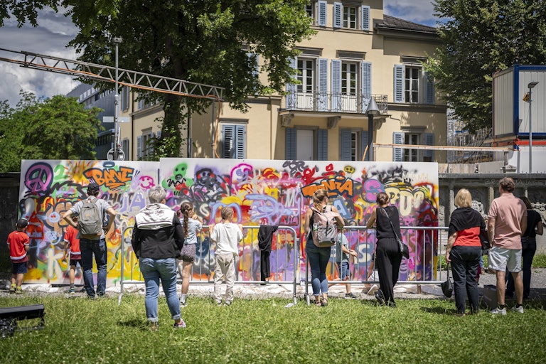 Menschen beobachten und fotografieren an einem sonnigen Tag eine lebendige Graffiti-Wand im Freien, hinter der sich ein Wohnhaus und Bäume befinden.