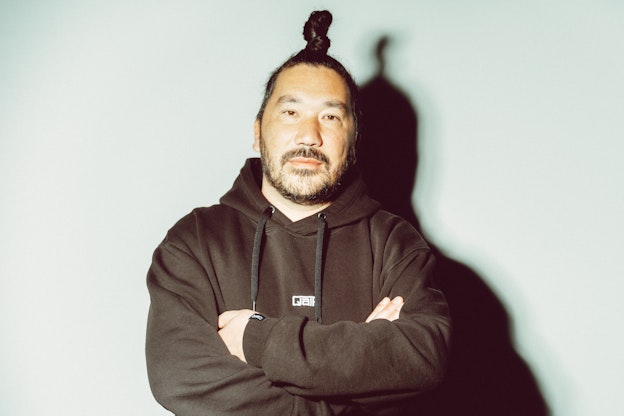 Ein Mann mit Haarknoten und Bart steht mit verschränkten Armen in einem schwarzen Kapuzenpulli vor einem hellen, schlichten Hintergrund.