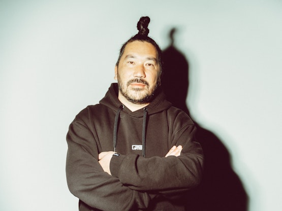 Ein Mann mit Haarknoten und Bart steht mit verschränkten Armen in einem schwarzen Kapuzenpulli vor einem hellen, schlichten Hintergrund.