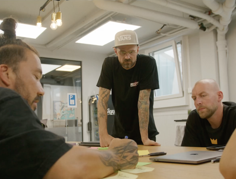 Drei tätowierte Männer sitzen in einem modernen Büro um einen Tisch herum, unterhalten sich und schreiben während einer Diskussion auf Klebezettel.