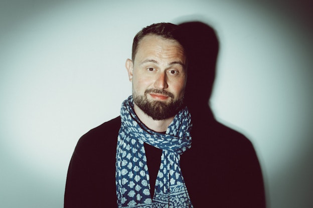 Ein bärtiger Mann mit einem gemusterten blauen Schal und einem schwarzen Hemd steht vor einem schlichten weißen Hintergrund, hinter dem sein Schatten liegt.