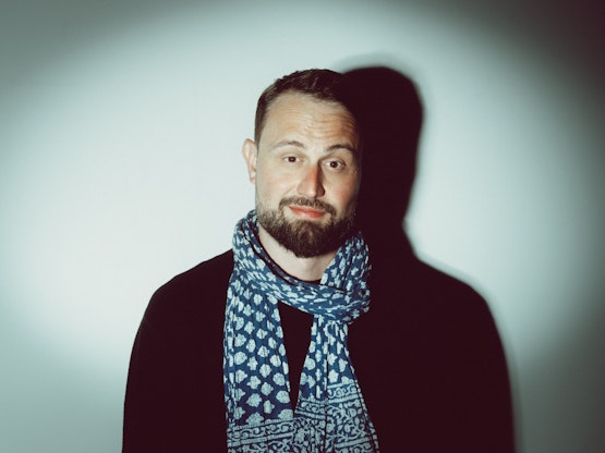 Ein bärtiger Mann mit einem gemusterten blauen Schal und einem schwarzen Hemd steht vor einem schlichten weißen Hintergrund, hinter dem sein Schatten liegt.