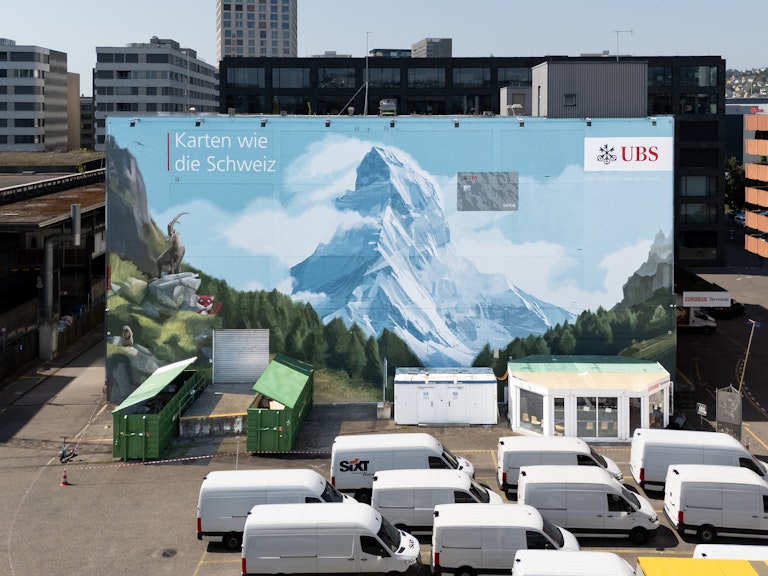 Grosse Wandmalerei des Matterhorns mit UBS-Logo und deutschem Text "Karten wie die Schweiz" auf einem Gebäude, daneben weisse Lieferwagen.
