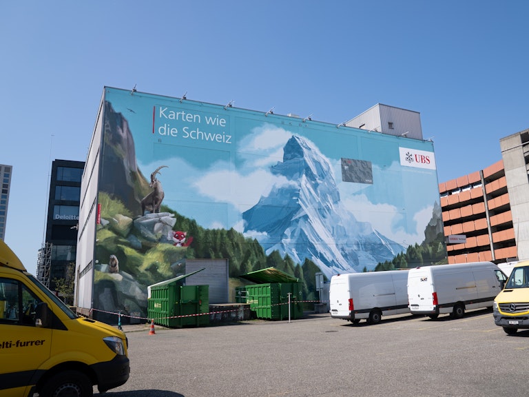 Ein grosses Wandbild zeigt Alpenberge und Steinböcke, das UBS-Logo und "Karten wie die Schweiz". Davor sind mehrere Autos geparkt.