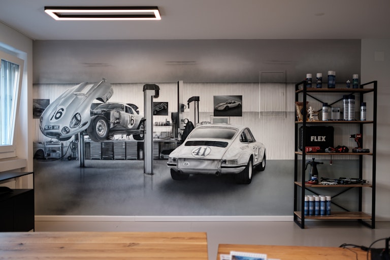 Ein Wandgemälde zeigt Oldtimer in einer Garage; daneben stehen Regale mit Autopflegeartikeln in einem aufgeräumten, modernen Raum mit Holz- und Metallakzenten.