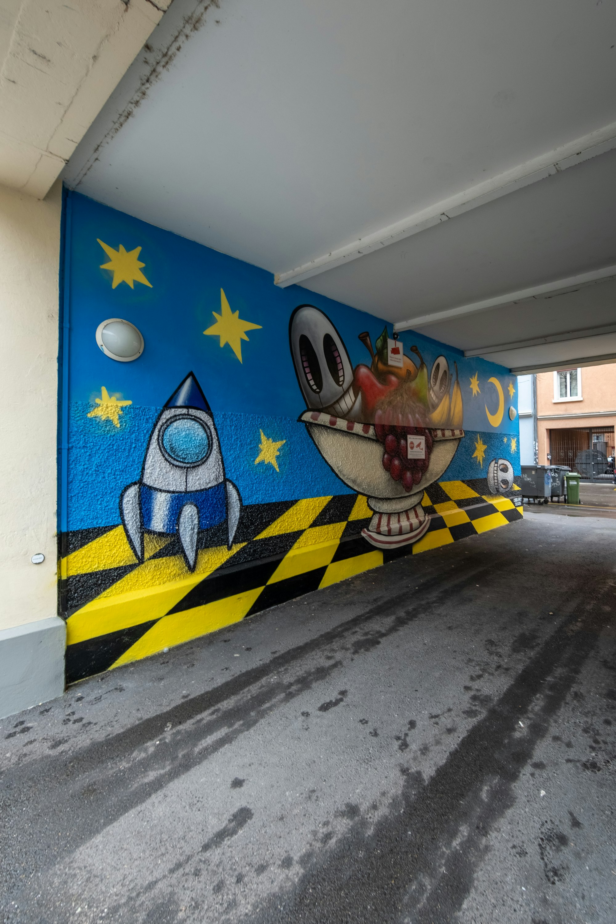 Ein Wandgemälde an einer blauen Wand zeigt eine Cartoon-Rakete, gelbe Sterne und eine Obstschale mit Augen über schwarz-gelben Streifen.