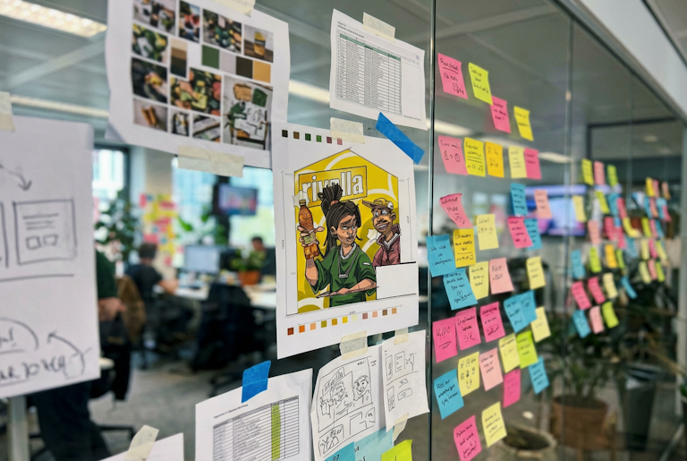 Eine Glaswand mit bunten Haftnotizen, Skizzen und Bildern in einem Büro, im Hintergrund Menschen, die an Schreibtischen arbeiten.