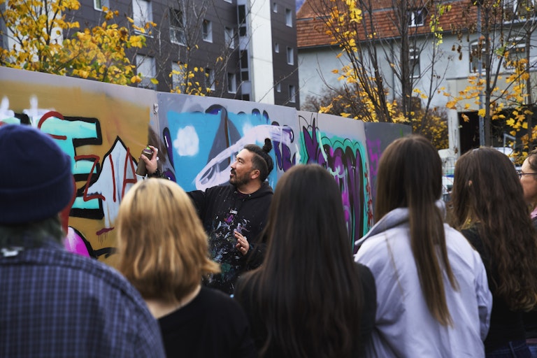 Ein Mann zeigt einer Gruppe in einem städtischen Gebiet im Herbst seine Graffitikünste an einer bemalten Wand.