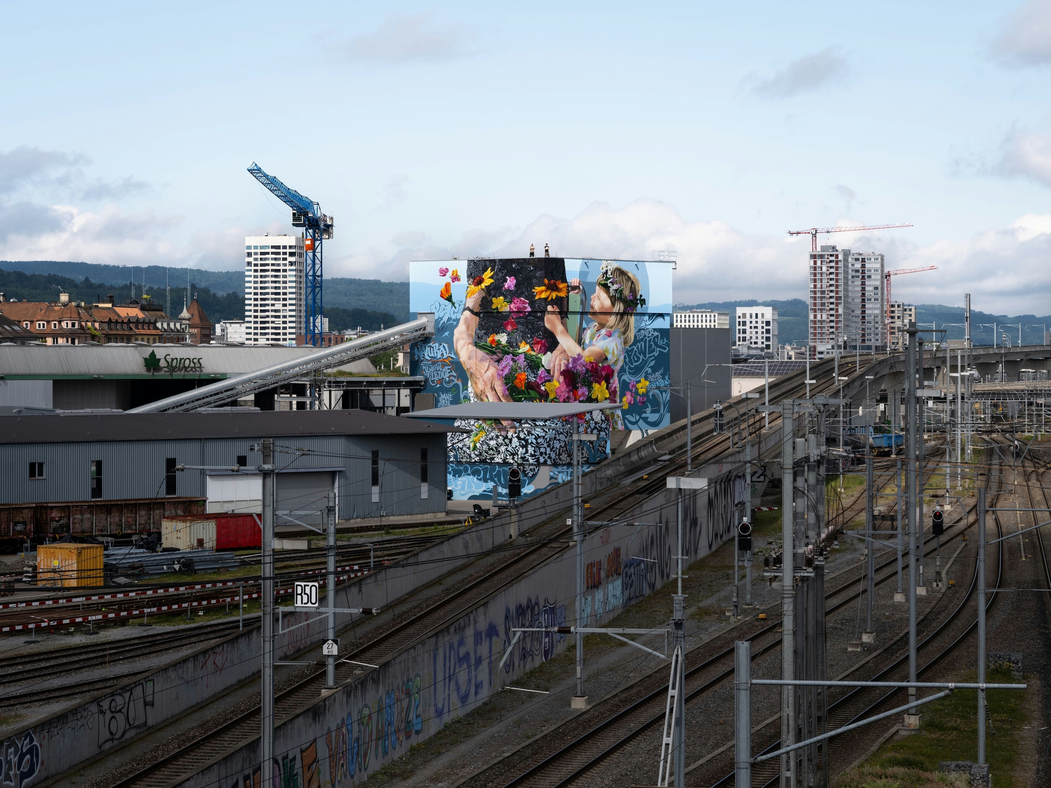 Ein lebhaftes Wandgemälde mit zwei Figuren schmückt ein Gebäude an den Bahngleisen in einem städtischen Gebiet mit Kränen und Hochhäusern dahinter.