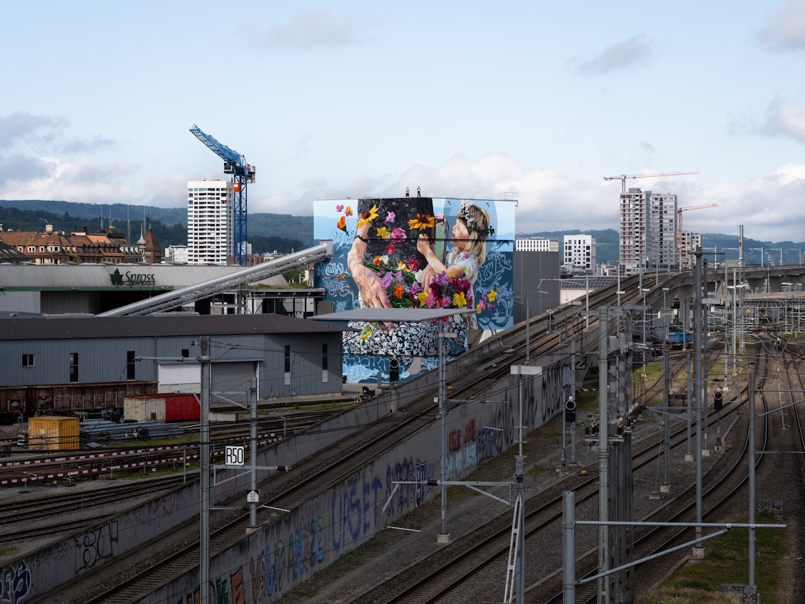 Ein lebhaftes Wandgemälde mit zwei Figuren schmückt ein Gebäude an den Bahngleisen in einem städtischen Gebiet mit Kränen und Hochhäusern dahinter.