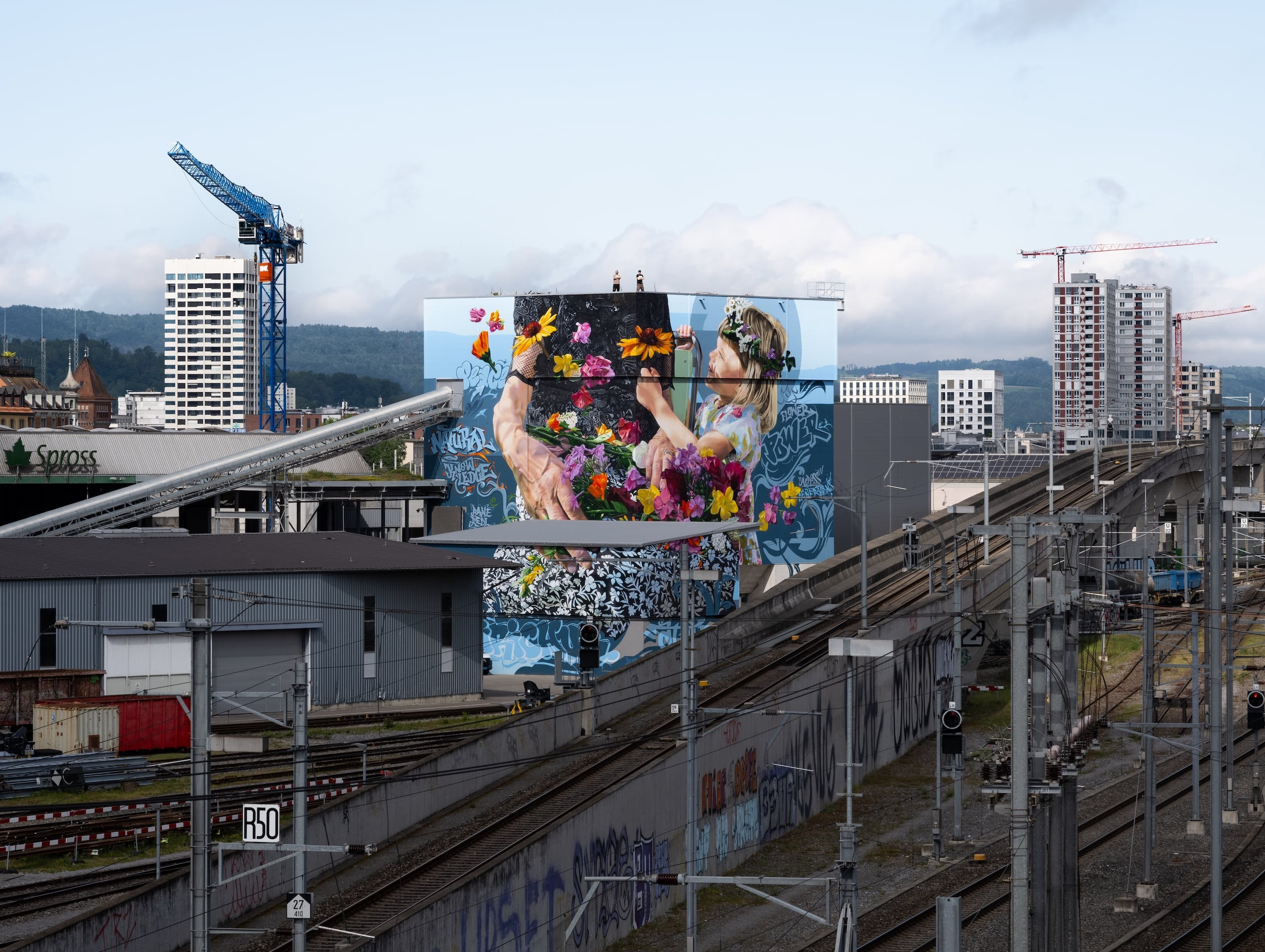 Ein lebhaftes Wandgemälde von zwei Menschen mit Blumen schmückt ein Gebäude neben Bahngleisen und Kränen in einem Industriegebiet.