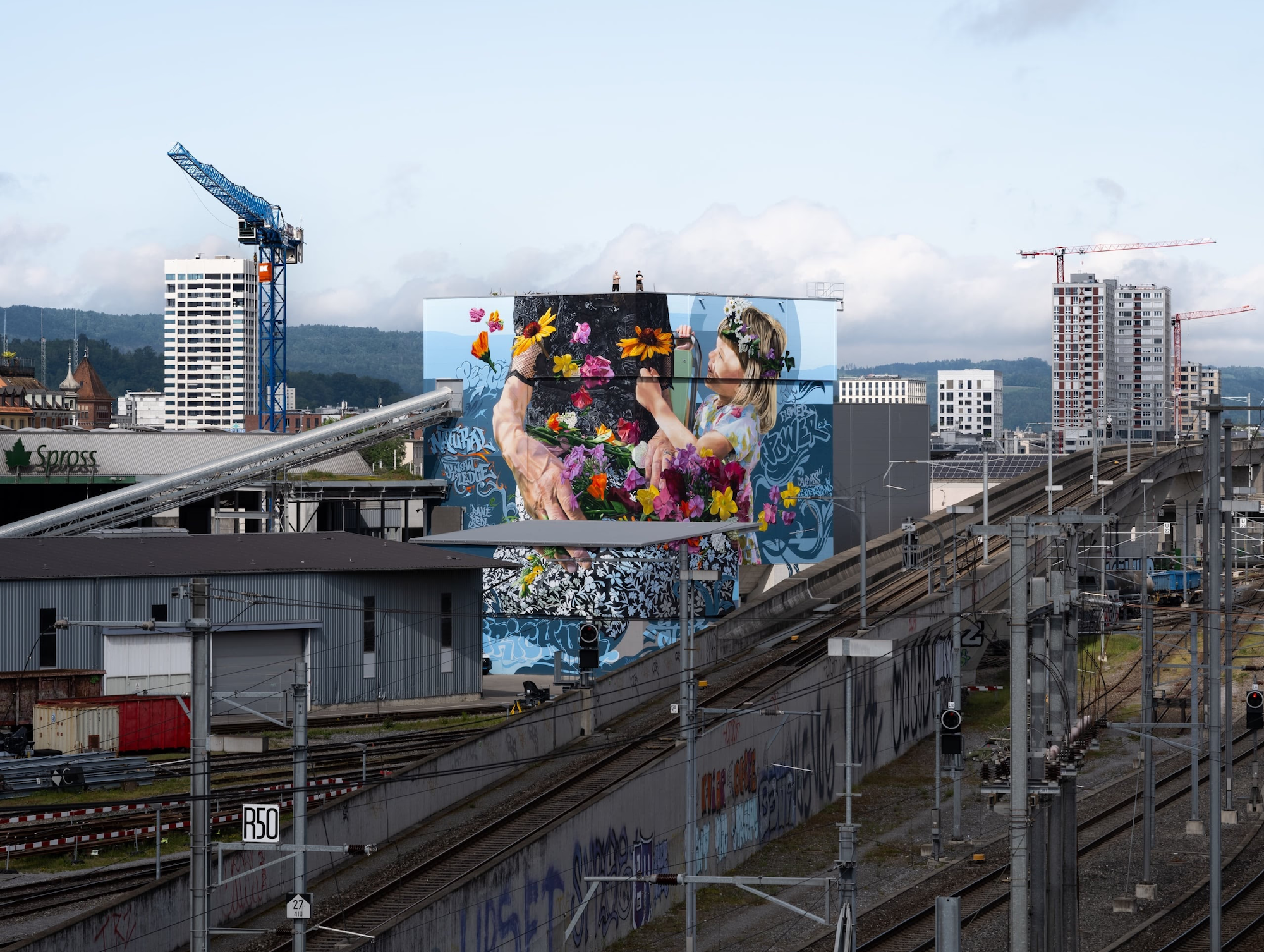 Ein lebhaftes Wandgemälde von zwei Menschen mit Blumen schmückt ein Gebäude neben Bahngleisen und Kränen in einem Industriegebiet.
