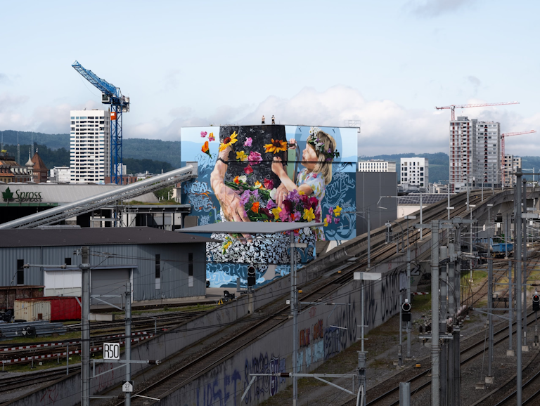 Ein lebhaftes Wandgemälde von zwei Menschen mit Blumen schmückt ein Gebäude neben Bahngleisen und Kränen in einem Industriegebiet.