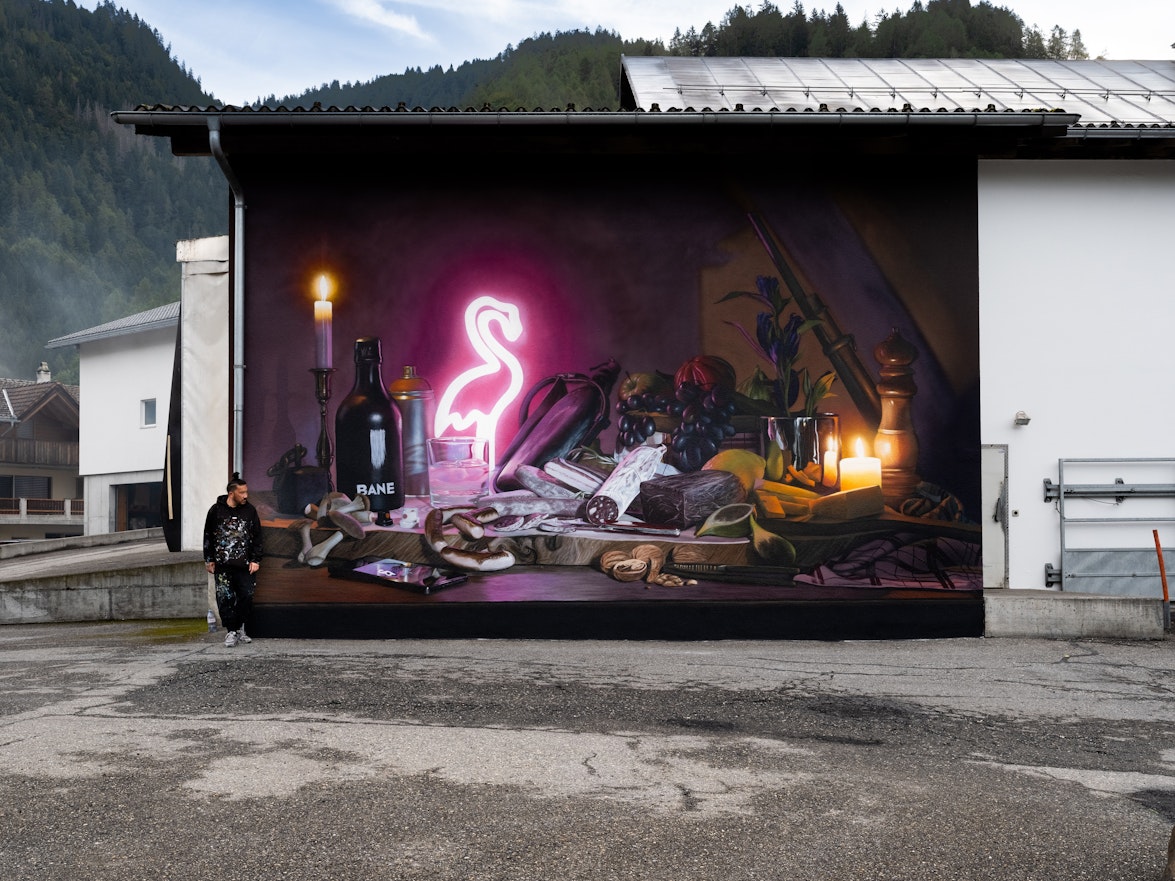 Eine Person steht neben einer Wandmalerei an einer Gebäudewand, die Kerzen, Früchte, Flaschen und einen leuchtenden rosa Neonflamingo zeigt.