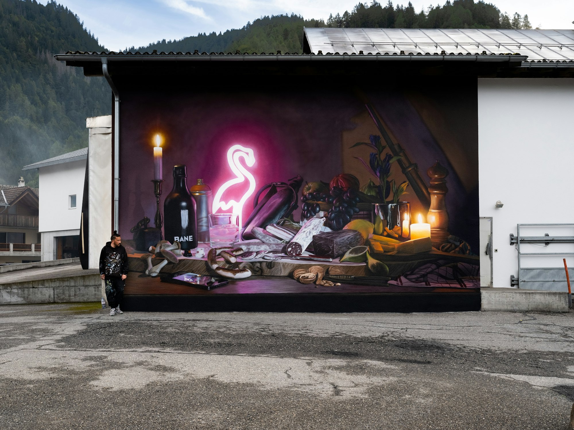 Eine Person steht neben einer Wandmalerei an einer Gebäudewand, die Kerzen, Früchte, Flaschen und einen leuchtenden rosa Neonflamingo zeigt.