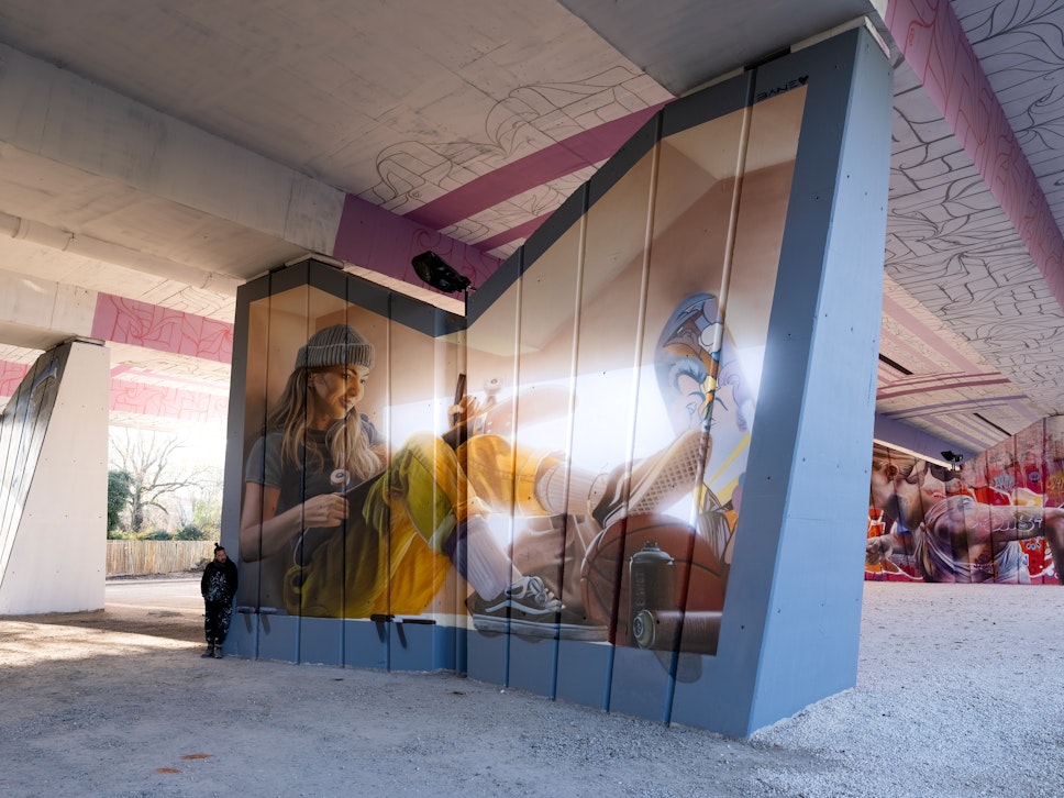 Eine Person steht in der Nähe eines großen Wandgemäldes unter einer Brücke, auf dem eine junge Frau mit Mütze und gelber Hose mit einem Skateboard sitzt.