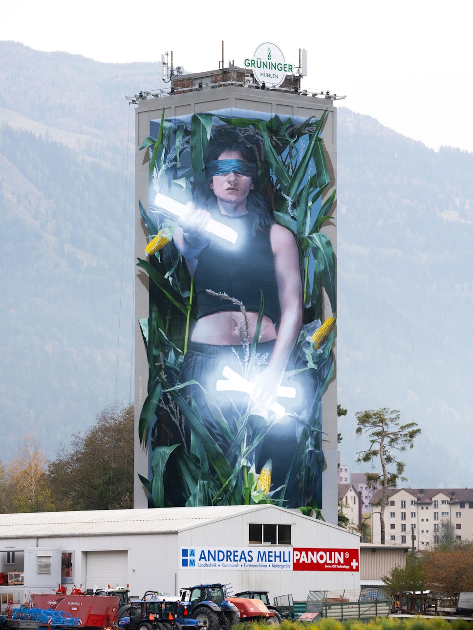 Ein hohes Gebäude zeigt ein Wandgemälde einer Frau mit verbundenen Augen und glühenden Stäben, umgeben von Maispflanzen und Firmenschildern am Sockel.