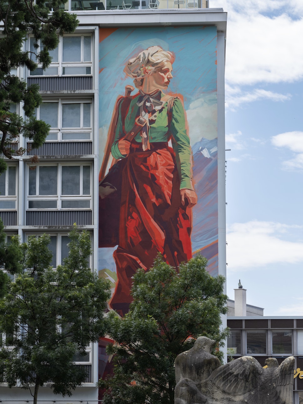Ein großes Wandgemälde mit einer Frau in grüner und roter Kleidung befindet sich auf einem modernen Gebäude, vor dem Bäume und ein Teil einer Skulptur stehen.