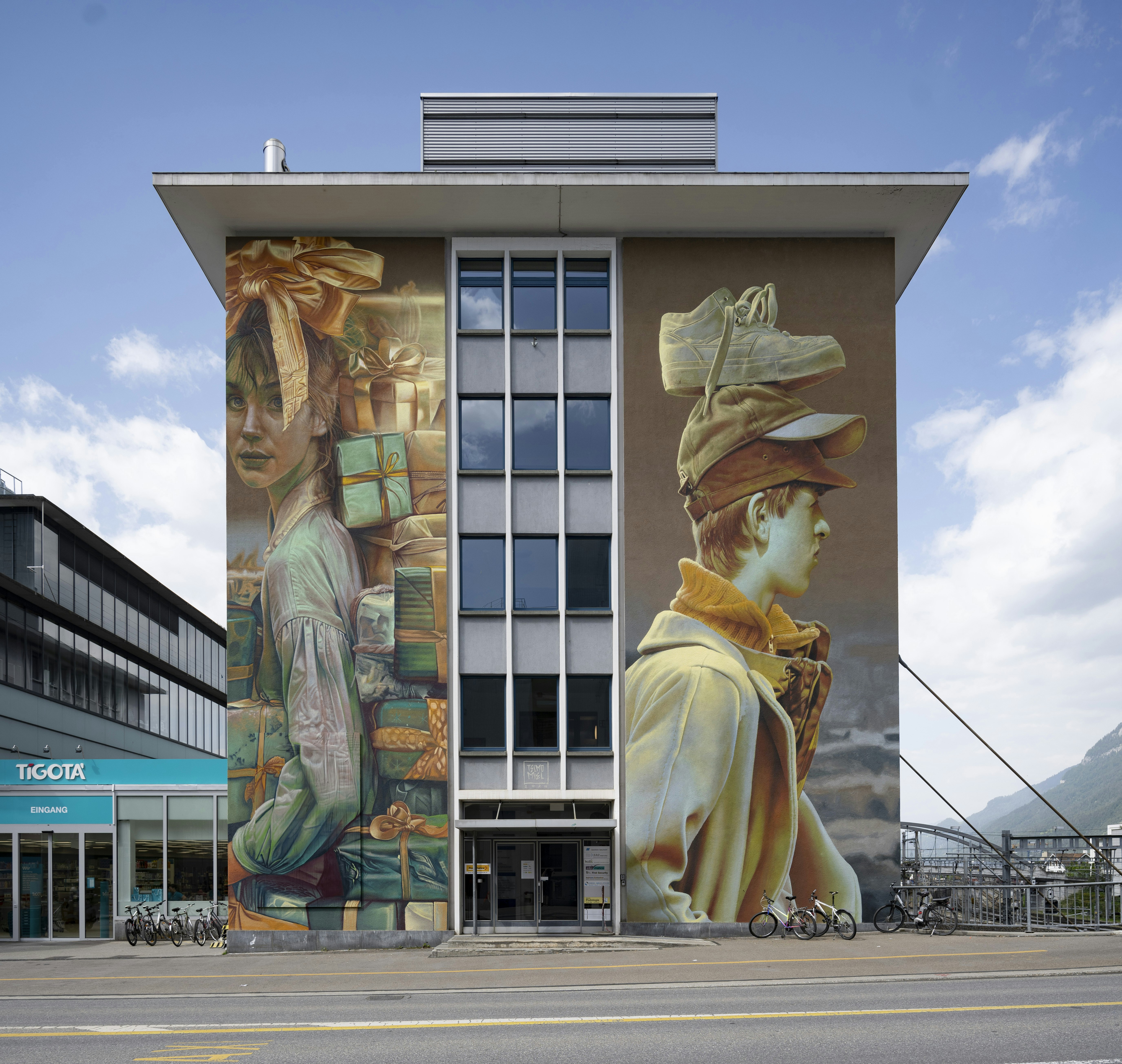 Modernes Gebäude mit zwei Wandmalereien: eine Person, die Geschenkkartons trägt, eine mit Schuhen auf dem Kopf. Unten sind Fahrräder geparkt.