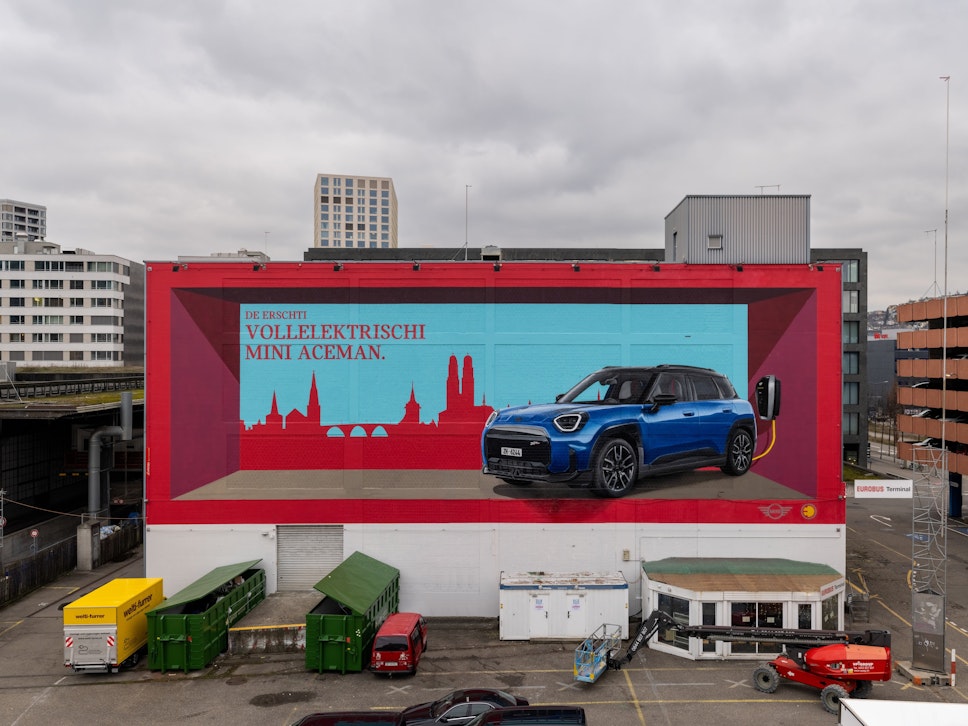 Ein Wandbild auf einem Gebäude zeigt einen blauen MINI Aceman EV, eine rote Stadtsilhouette und einen deutschen Text. Davor sind mehrere Autos geparkt.