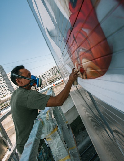 Artist Alex Hohl mit Atemschutzmaske sprüht ein Wandbild auf die Aussenwand von Spross Recycling in Hardbrücke, Zürich, während er auf einem Gerüst steht.