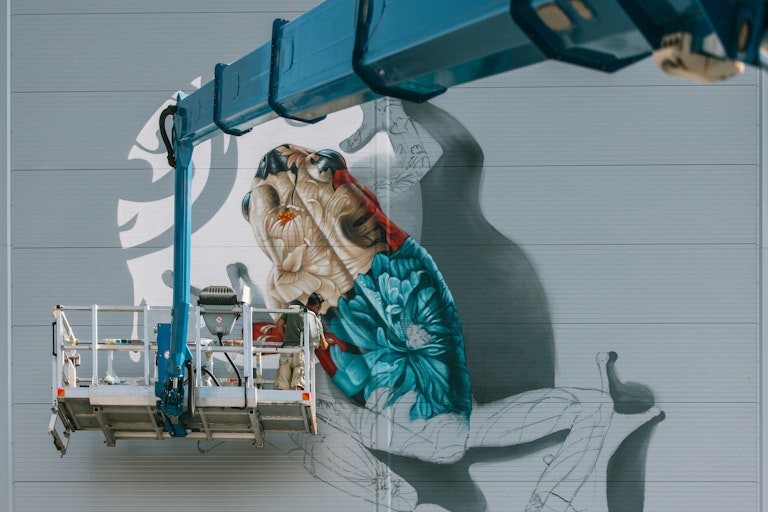 Artist Alex Hohl steht auf einer Hebebühne und malt ein Wandgemälde an die Fassade von Spross Recycling in Zürich, CH.