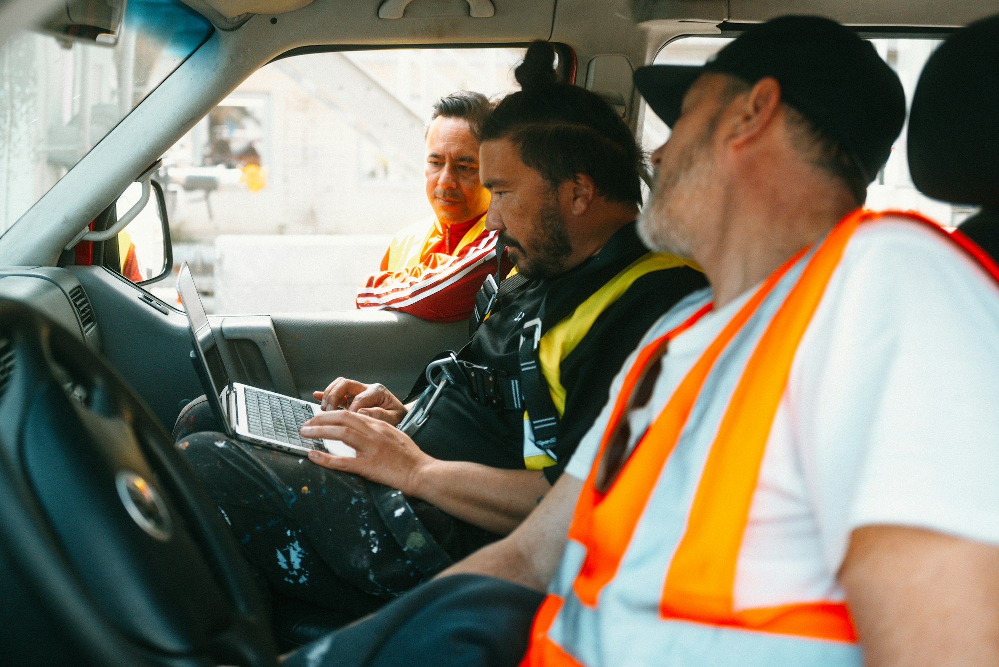 Drei Männer in Sicherheitswesten sitzen in einem Fahrzeug; einer arbeitet mit einem Laptop, während die anderen aufmerksam zuhören und beobachten.