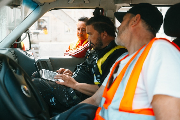 Drei Männer in Sicherheitswesten sitzen in einem Fahrzeug; einer arbeitet mit einem Laptop, während die anderen aufmerksam zuhören und beobachten.
