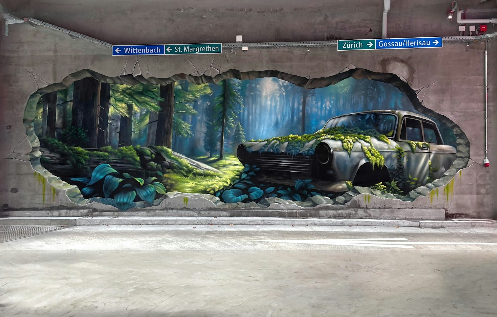 Eine Wandmalerei mit einer Waldszene und einem moosbewachsenen alten Auto ziert eine Parkhauswand, die wie aufgesprengt wirkt. Strassenschilder oben.