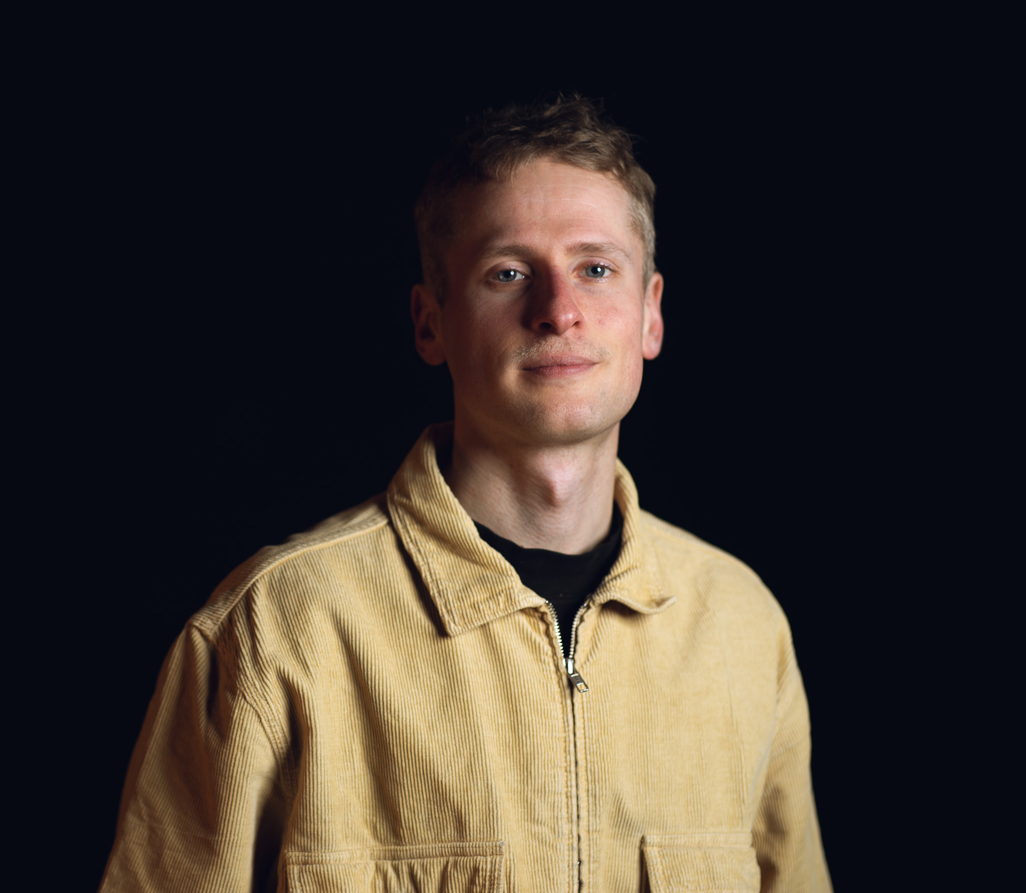 Eine kurzhaarige Person in einer beigen Kordjacke steht vor einem dunklen Hintergrund und blickt mit neutralem Blick in die Kamera.