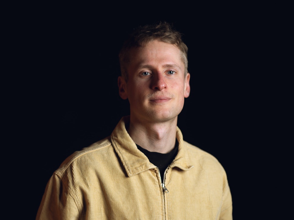 Eine kurzhaarige Person in einer beigen Kordjacke steht vor einem dunklen Hintergrund und blickt mit neutralem Blick in die Kamera.