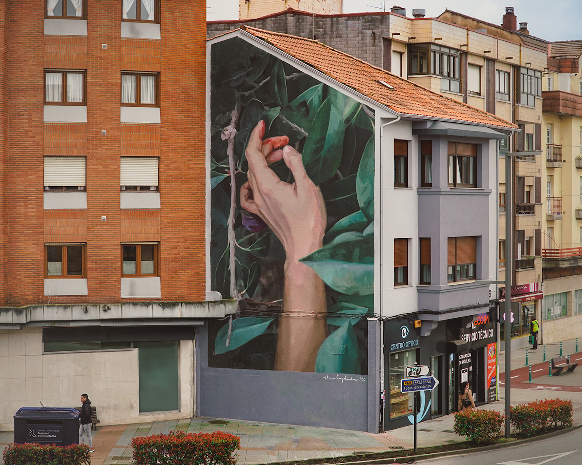 Ein Wandbild der WAND AG zeigt eine Hand, die eine rote Blume hält, auf einem Gebäude neben belebten Strassen, Geschäften und Wohnungen in der Stadt.
