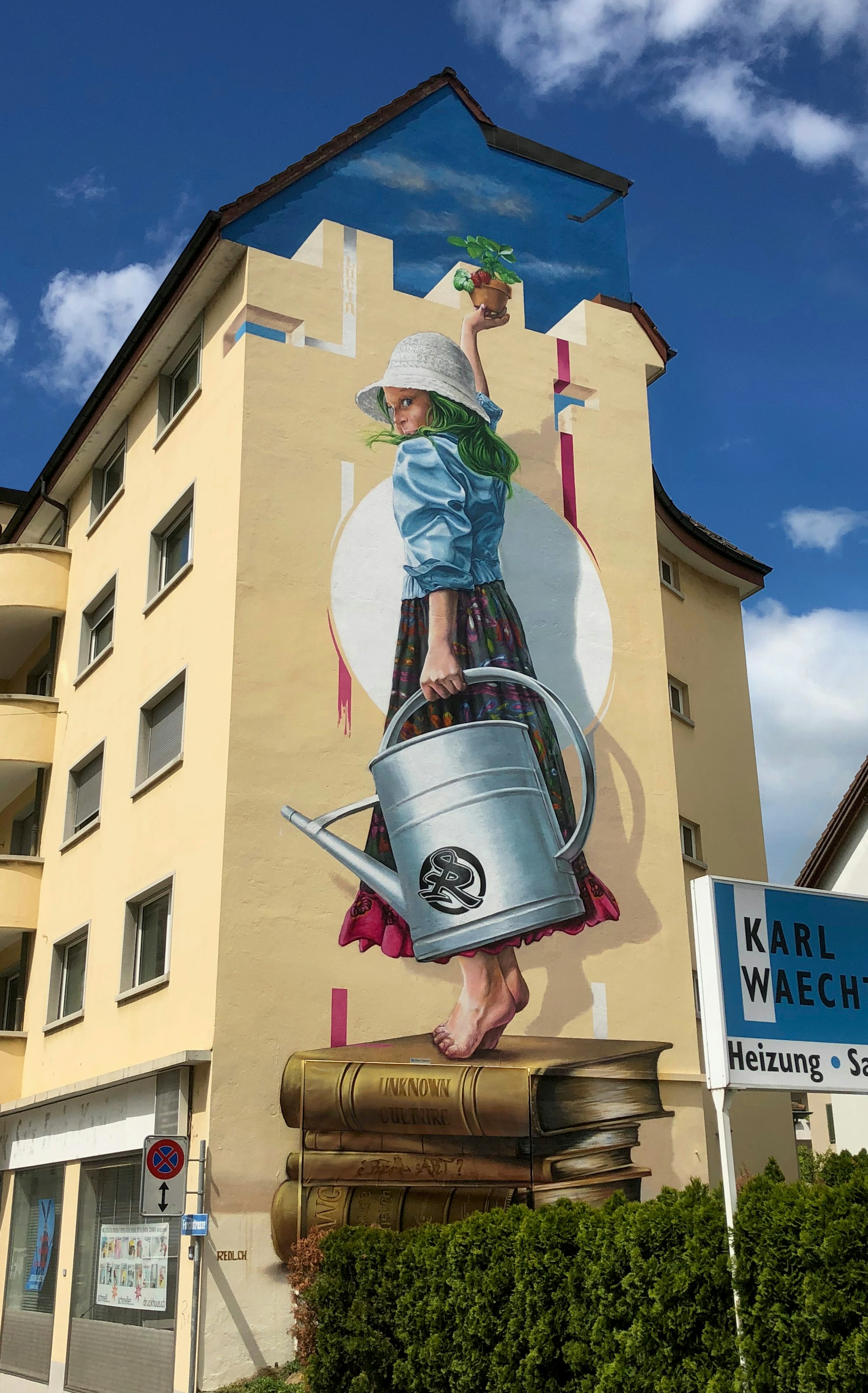 Ein Wandbild der WAND AG zeigt ein grünhaariges Mädchen auf Büchern, das eine Pflanze und eine Giesskanne unter einem blaün Himmel mit Wolken hält.