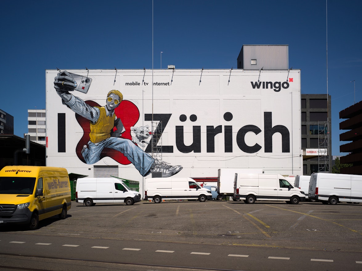 Das Wandbild der WAND AG mit "I ❤️ Zürich" und einer Karikatur mit Boombox; davor stehen weisse Lieferwagen und ein gelber Lieferwagen.
