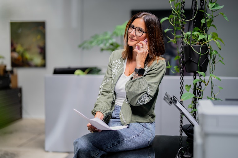 Frau mit Brille und grüner Jacke in einem Haus, hält Papiere in der Hand, telefoniert; hinter ihr Pflanzen in einer kreativen, Street-Art-ähnlichen Szene.