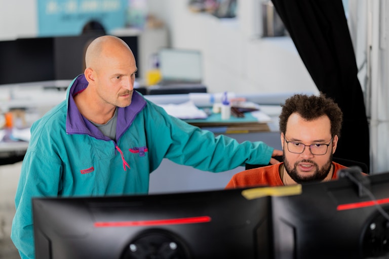 Zwei Männer arbeiten in einem Büro an einem Computer. Der eine legt dem sitzenden Mann die Hand auf die Schulter, während sie auf den Monitor schaün.