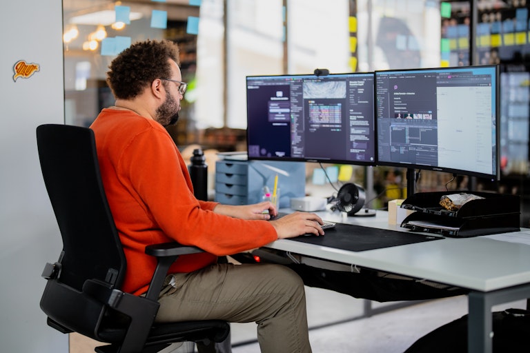 Ein Mann in einem orangefarbenen Pullover arbeitet an einem Schreibtisch mit zwei grossen Monitoren, auf denen Code und Projektwerkzeuge in einem modernen Büro angezeigt werden.