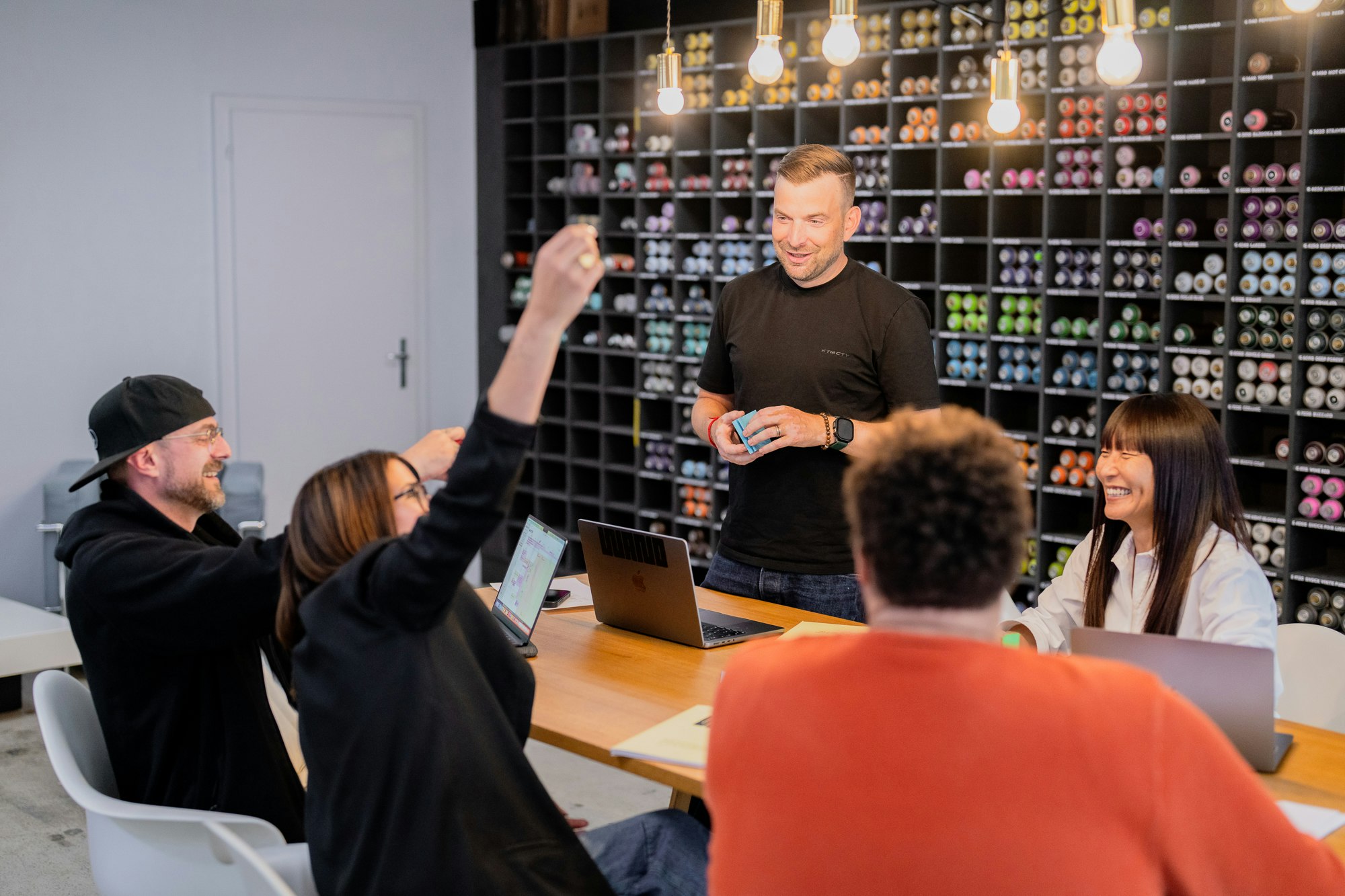 Fünf Personen an einem Tisch mit Laptops, einer hebt aufgeregt die Faust während einer Besprechung; im Hintergrund sind bunte Regale zu sehen.