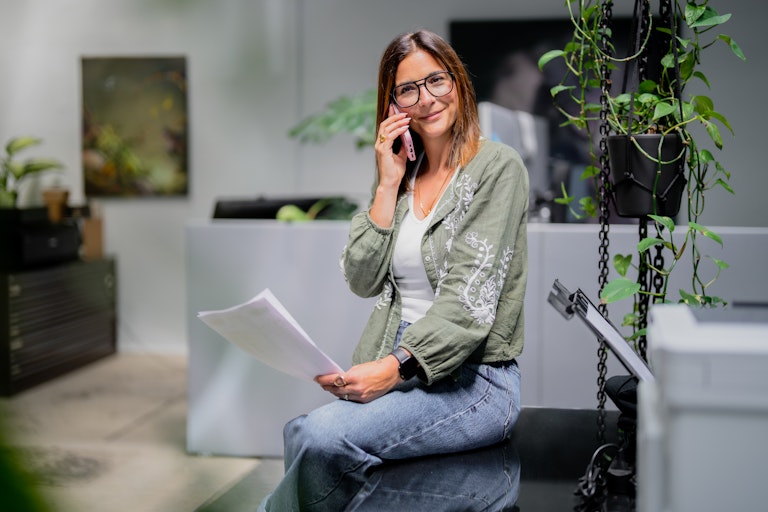 Eine Frau mit Brille sitzt an einem Schreibtisch, hält Papiere in der Hand und spricht mit ihrem Smartphone in einem modernen Büro mit Pflanzen.