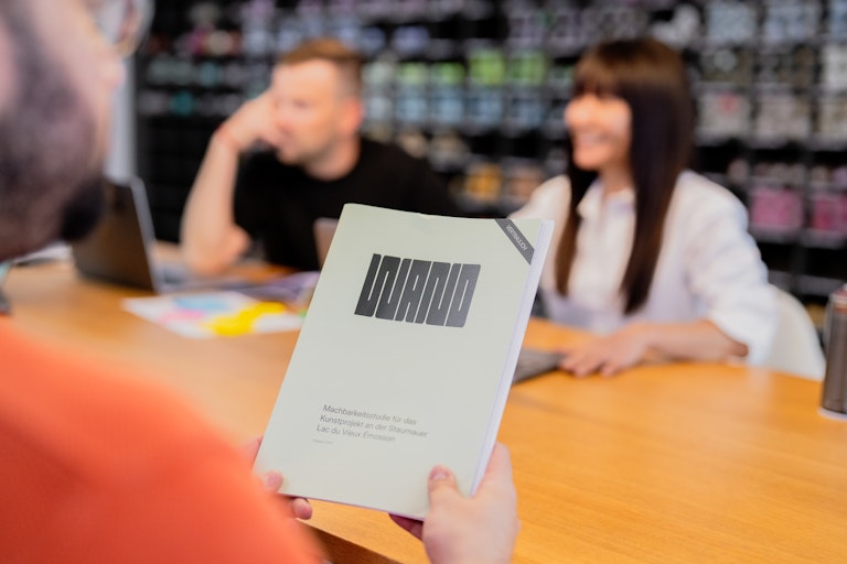 Person mit einer Broschüre an einem Tisch; zwei weitere Personen im Hintergrund diskutieren an Laptops in einem hellen, modernen Arbeitsraum.