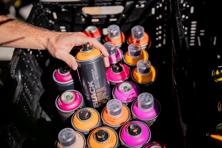 Eine Hand hebt eine schwarze Sprühdose aus einer Kiste mit bunten Dosen in Rosa, Orange und Gelb auf.