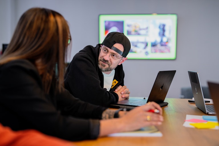 Zwei Menschen mit Laptops unterhalten sich an einem Tisch; einer trägt eine Mütze nach hinten. Ein Bildschirm hinter ihnen zeigt bunte Bilder.
