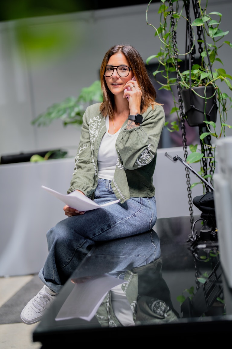 Eine Frau mit Brille, grüner Jacke und Jeans sitzt auf einem Glastisch, hält Papiere in der Hand und telefoniert in einem mit Pflanzen gefüllten Raum.