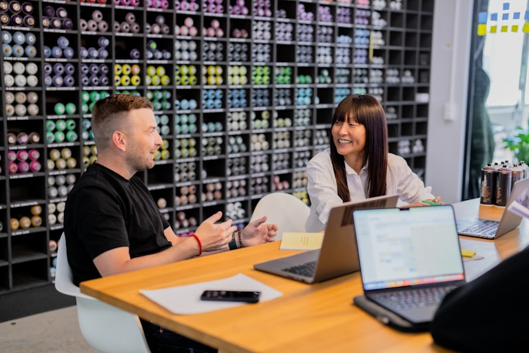 Zwei Personen sitzen an einem Tisch mit Laptops und Papieren und unterhalten sich, während im Hintergrund Regale mit bunten Sprühdosen zu sehen sind.