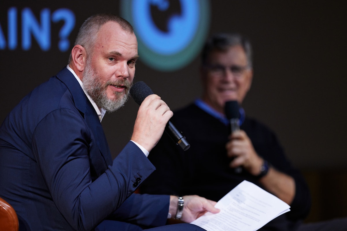 A man in a blue suit speaks into a microphone while holding papers, sitting next to another man in glasses who is also holding a microphone, during a discussion or interview event.