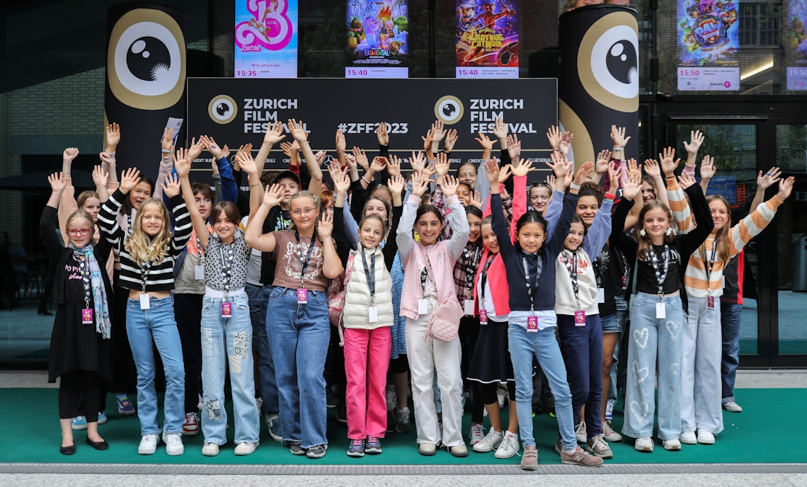 A large group of smiling teenagers with raised hands pose together outside at the Zurich Film Festival, in front of festival signage and posters.