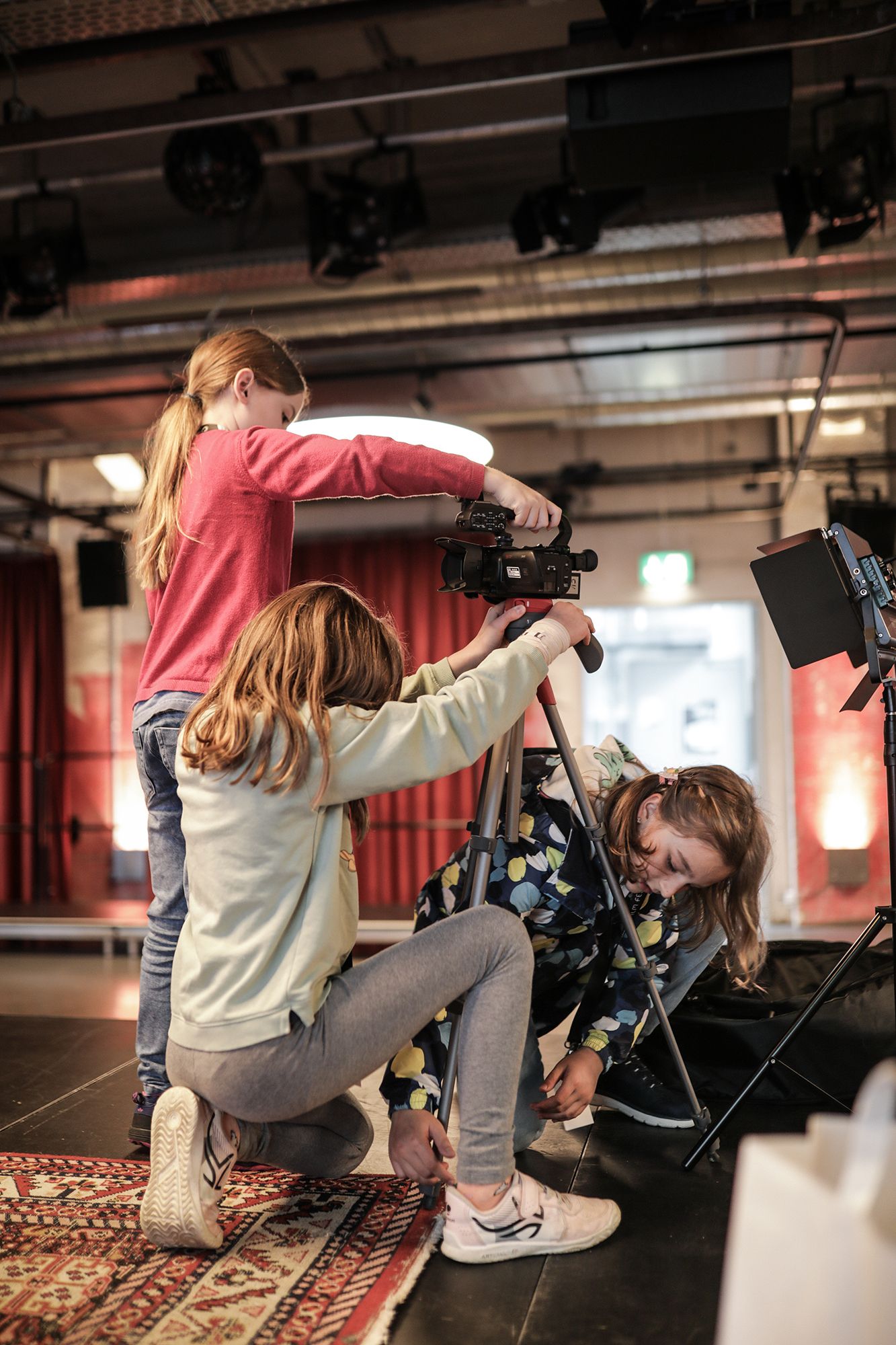 Drei Kinder stellen im Haus eine Videokamera auf einem Stativ auf. Ein Mädchen richtet die Kamera aus, ein anderes hält das Stativ, und ein drittes kniet und befestigt ein Stativbein. Sie wirken konzentriert und arbeiten zusammen.
