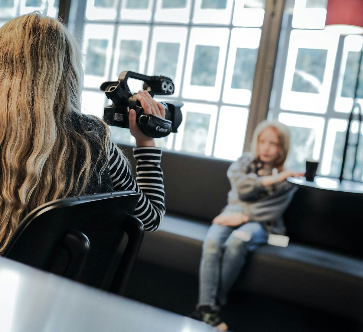 Eine Person mit langen, welligen blonden Haaren filmt ein kleines Kind, das auf einer gepolsterten Bank vor einem großen Fenster sitzt. Das Kind gestikuliert beim Sprechen mit einer Hand.